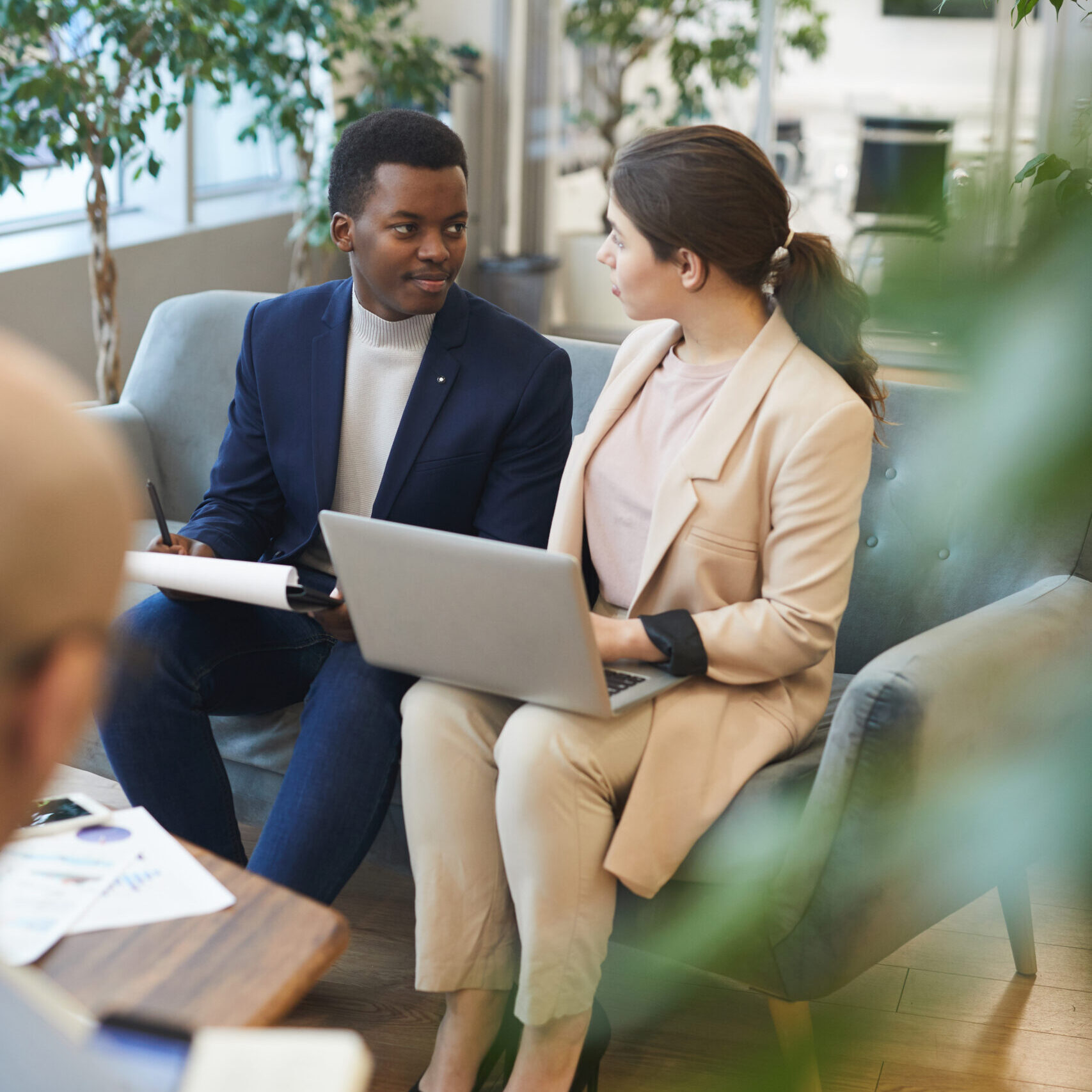 High angle portrait of successful African businessman talking to female partner while working in comfortable chair at office building hall decorated with plants, copy space